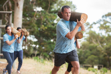 Determined man running with log on boot camp obstacle course - obrazy, fototapety, plakaty