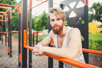 Obraz premium Portrait young bearded man standing on public sports ground and training on parallel bars. Handsome workout athlete. Portrait sporty man with beard, rest after training. Summer workout outdoors