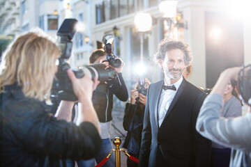 Smiling celebrity in tuxedo being photographed by paparazzi at red carpet event