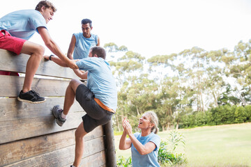 Teammates helping man over wall on boot camp obstacle course - obrazy, fototapety, plakaty