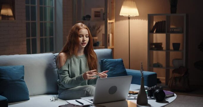 Pretty caucasian redhead girl sitting on coach looking at laptop, having a videochat or online tutorial, recording an internet vlog, cute model looking at webcamera and talking 4k footage