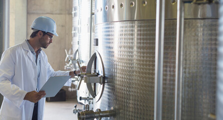 Vintner in lab coat hard hat examining stainless steel vat in cellar