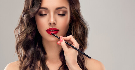 Makeup artist applies  red lipstick  . Beautiful woman face. Hand of visagiste, painting  cosmetics of young beauty  model girl . Make up in process - obrazy, fototapety, plakaty