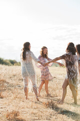 Boho women holding hands and dancing in circle in sunny rural field