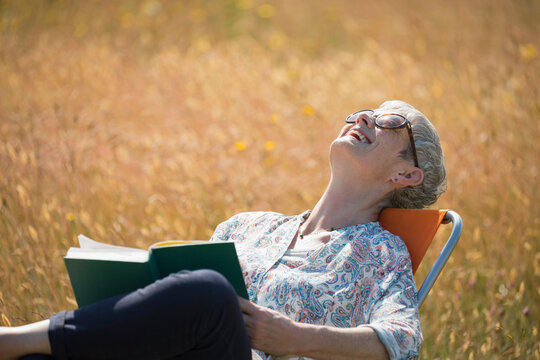 Senior Woman Reading Book And Laughing With Head Back In Sunny Field