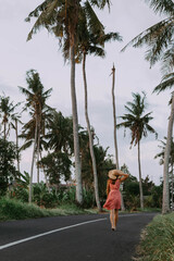 A young beautiful woman in a dress and hat walks along the road with palm trees