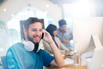 Fototapeta premium Smiling creative businessman with headphones talking on cell phone in office