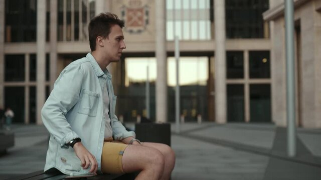 Young man in white shirt and bright jacket  takes off his medical mask and put it on brench near modern office during pandemic covid-19 coronavirus quarantine.
