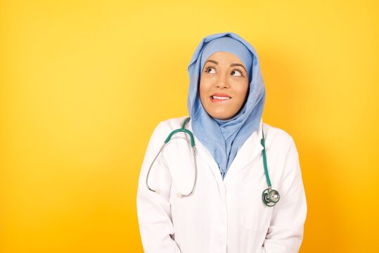 Photo of amazed Young beautiful doctor muslim woman wearing a medical uniform and hiyab bitting lip and looking up to empty space, isolated over background.