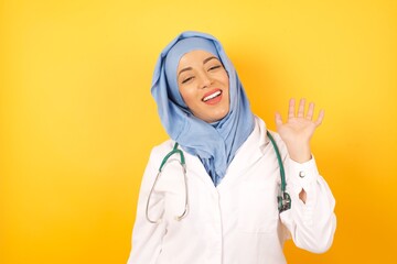 Young beautiful doctor muslim woman wearing a medical uniform and hiyab, over yellow isolated background Waiving saying hello happy and smiling, friendly welcome gesture.