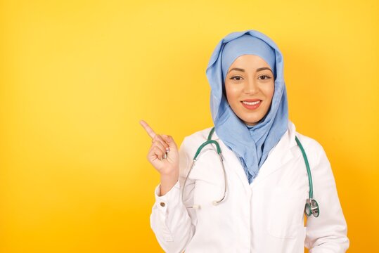 Smiling Doctor Muslim Woman Wearing Medical Uniform Indicating Finger Empty Space Sales Manager Wearing Medical Uniform And A Stethoscope Isolated Yellow Color Background