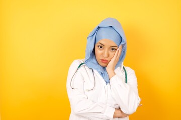 Very bored adult doctor woman wearing medical uniform and hiyab holding hand on cheek while support it with another crossed hand, looking tired and sick, over yellow background.