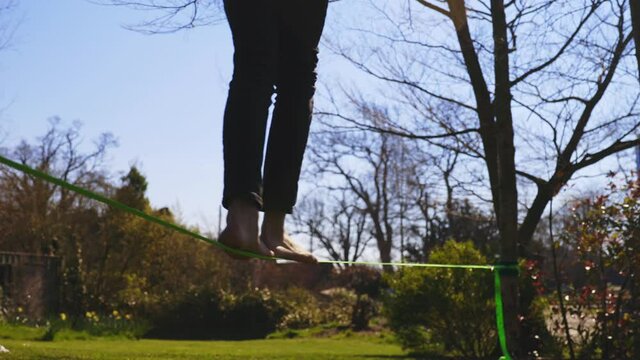 Low angle of young barefoot man wearing black jeans and puffer coat walking on, then falling off slack line. Trees with no leaves and green bushes and grass in background.