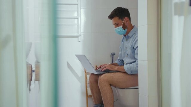 Happy businessman working on laptop wearing mask sit on the toilet at home restroom bearded funny wc isolation quarantine pandemic coronavirus covid-19 portrait close up slow motion