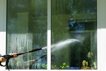 A woman washes a window with a spray under high pressure. Indoor plants are visible through the glas