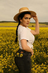 beautiful woman with dark hair in casual clothes in straw hat posing in blooming rapeseed field