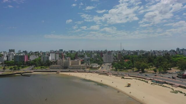 Aerial shot of people at beach by street in city against blue sky, drone flying forward over sea on sunny day - Montevideo, Uruguay