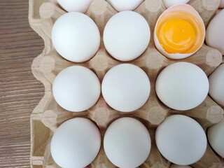 White eggs carton & cracked egg half with yolk top view on wooden background. Broken and whole eggs in carton box. Poultry eggs in carton paper tray package - healthy food breakfast cooking concept