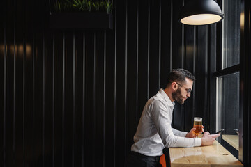 Side view of attractive bearded man standing in bar after work, having glass of beer and using smart phone for hanging on social media.