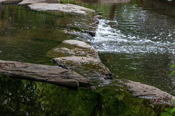 Calm floating creek with crystal clear water and rocks of a natural little dam or adventure hiking trail over a flat little brook in the woods as adventure tour for tourism with environmental health
