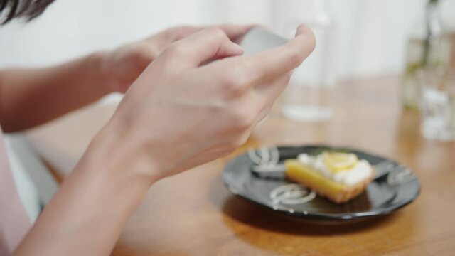 Close up of Asian woman hands using a smartphone to take a photo of lemon cake in a restaurant. Young woman relax sitting at the table and taking a photo of dessert in front of him in the cafe.
