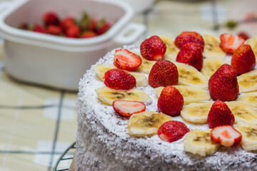 Homemade fruit cake with cream. Cake decorated with strawberries and bananas. Sprinkled with coconut on the sides. Cake on the right side of the screen. In the background is a cup with strawberries.