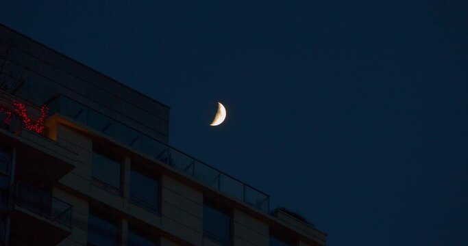 Lockdown time lapse shot of half moon over building in blue sky at night - Vancouver, Canada