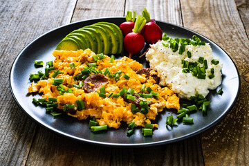 Delicious breakfast - scrambled eggs with fried sausages, cottage cheese, and avocado served on black plate on wooden table
