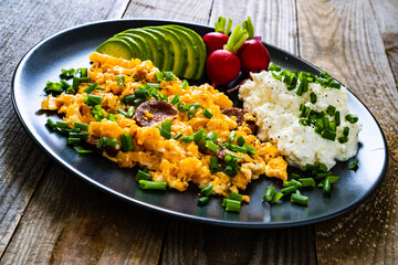 Delicious breakfast - scrambled eggs with fried sausages, cottage cheese, and avocado served on black plate on wooden table
