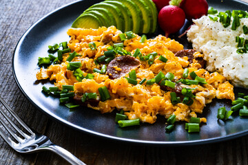 Delicious breakfast - scrambled eggs with fried sausages, cottage cheese, and avocado served on black plate on wooden table
