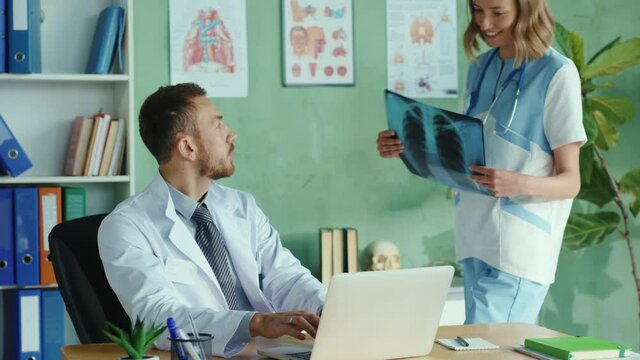 Serious caucasian doctor working at the office on portable laptop. Daring young nurse flirting with therapist trying to seduce him. Sexual harrasment in the hospital. Problems.