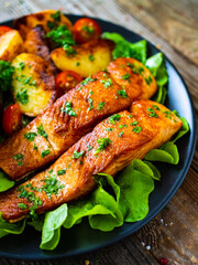 Fried salmon steak with potatoes and vegetables served on black plate on wooden table