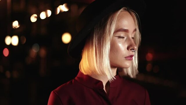 Portrait of trendy hipster woman with blond hairstyle standing on city street at night. Hat, nose piercing. Mysterious girl. Slow motion.