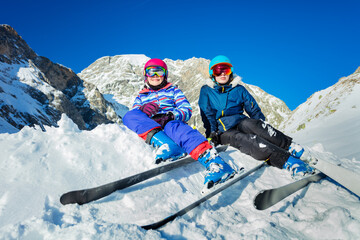 Two smiling happy girls with mountain ski attached sit on the pile of snow in colorful sport outfit helmet and masks