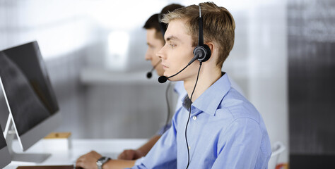 Two young men in headset, sitting at the desk in the modern office, listening to the clients. Call center operators at work