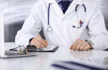 Fototapeta premium Unknown male doctor sitting and working with clipboard of medication history record in clinic at his working place, close-up. Young physician at work. Perfect medical service, medicine concept