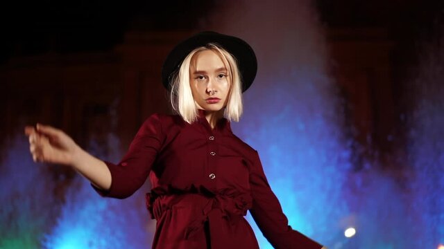 Portrait of millennial hipster woman with blond hairstyle standing on neon fountain background. City at night. Hat, nose piercing. Beautiful attractive girl. Slow motion.