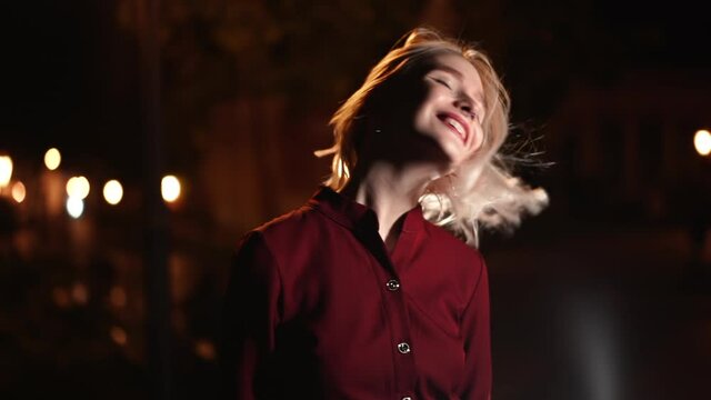 Portrait of millennial hipster woman with blond hairstyle makes funny faces, fooling around. City street at night. Hat, nose piercing. Funny girl. Slow motion.