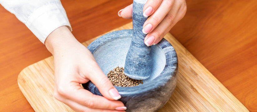 Hands Of Woman Grinding Black Pepper By Pestle In A Gray Granite Mortar On The Table.