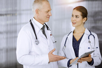 Elderly confident doctor and woman-physician discussing current disease therapy while standing in sunny clinic. Teamwork in medicine