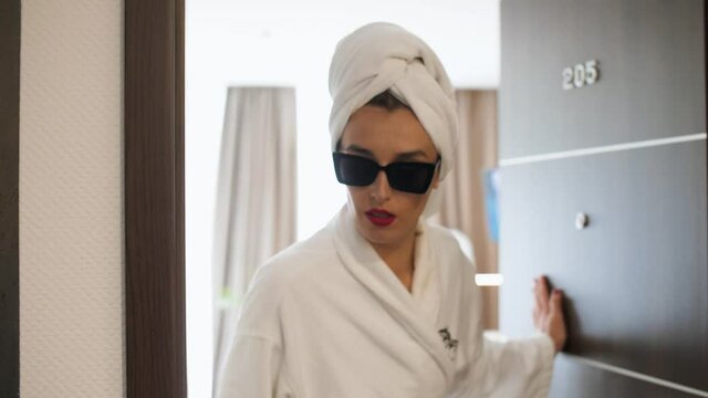 a girl in a white bathrobe in a bone room with a towel on her head with red earrings and sunglasses, opens the number 205, looks out from side to side and closes the door