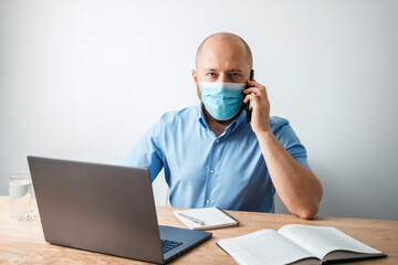 Bald business man with beard wearing surgical medical mask on face as protection against coronavirus, covid-19, talking on the phone, working or studying online on laptop at office