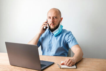 Bald business man with beard put off surgical medical mask as protection against coronavirus, covid-19 and talking on the phone, modern laptop on wooden table in office, white wall as background