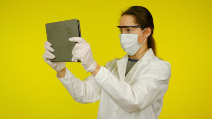 Young doctor in a medical mask, goggles and latex gloves looks at the tablet. Girl in a white coat on a yellow background holds a tablet in hands and looks at patient information. Coronavirus, flu