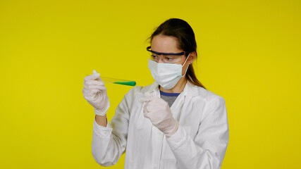 Doctor in a medical mask, goggles and latex gloves looks at the tests in the tube. Young girl in a white coat on yellow background holds hand a tube with green liquid