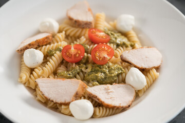 fussili pasta with chicken fillet, tomatoes, mozzarella and pesto sauce in white bowl closeup