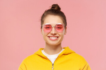 Close-up portrait of young smiling girl wearing yellow casual hoodie and round colored eyeglasses, hair tied in bun, isolated on pink background