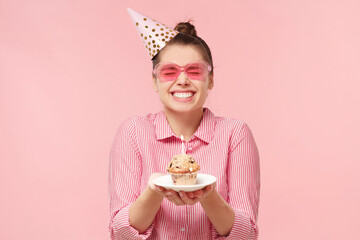 Excited funny teenage girl wearing colored glasses and birthday hat, holding cupcake on plate with...