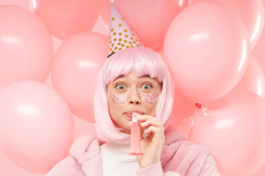 Headshot Portrait Of Young Beautiful Female Wearing Winter Bomber, Wig And Birthday Hat, Blowing Whistle Among Balloons, Isolated On Pink Background