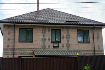 Naklejka premium facade of a brown private brick house with a row of windows under a tiled roof behind a metal fence against a gray sky